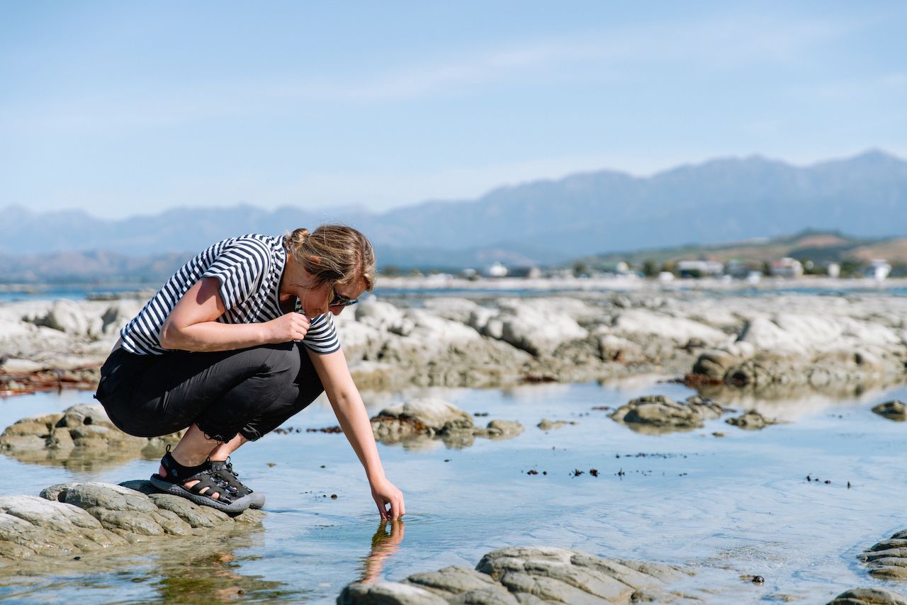 Eine Frau, die KEEN Outdoor Sandalen trägt, hockt auf einem Stein am flachen Wasser, eventuell am Meer oder an einem See, und greift mit der Hand ins Wasser.
